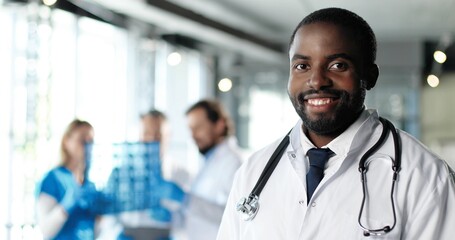Portrait of African American young man physician in white gown and stethoscope looking at camera and smiling in clinic. Handsome male doctor in hospital. Doctors on background. Healthcare.