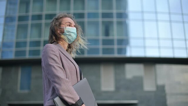 A young business woman in a medical mask and with a folder of documents stands against the background of a business center and looks meaningfully into the distance. The need to wear a mask due to an i