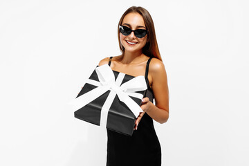 Beautiful young woman in black dress and sunglasses standing on a white background with a gift in her hands, Black Friday concept, shopping