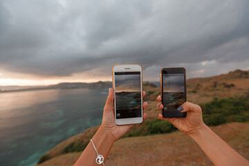  Two women tourist taking photo on sea beach with mobile smart phone camera at sunset, hands close up. Meresse hill Lombok Indonesia