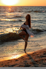 Fototapeta na wymiar Young beautiful woman in black bikini and white shirt on a tanned body is dancing on the beach at sunset. Soft selective focus, art nose.