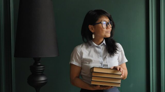 Professional model with long loose hair and glasses poses near black lamp and green wall holding books and smiling at home slow motion