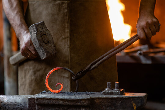 Old blacksmith is processing a hot metal object of a spiral shape on the anvil in the forge