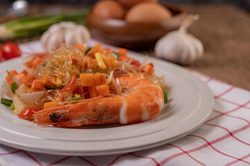 Stir-fried prawns with glass noodles in a white plate placed on a cloth with eggs and garlic.
