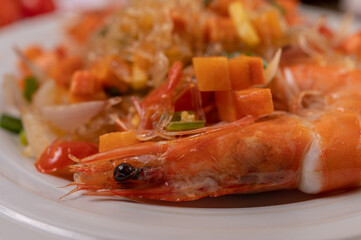 Stir-fried prawns with glass noodles in a white plate placed on a cloth with eggs and garlic.