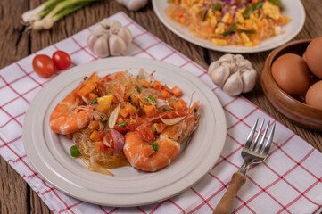 Stir-fried prawns with glass noodles in a white plate placed on a cloth with eggs and garlic.