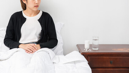 An uncomfortable woman sits on the bed and has medicine on the table.