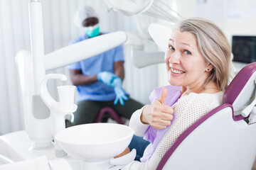 Happy cute mature woman sitting in dental chair after teeth cure giving thumb up. High quality photo