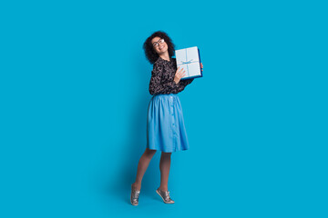 Fototapeta premium Monochrome photo of a caucasian woman with curly hair and glasses posing in a dress on a blue wall holding a present