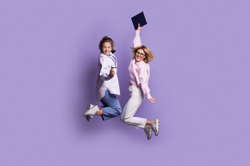 Two caucasian students in casual clothes are jumping on a violet studio wall while holding a book and smile at camera