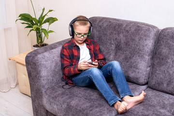 Obraz premium A charming 10-year-old boy in jeans and a checkered red shirt, wearing glasses and large headphones is listening to music or listening to a lesson on his smartphone