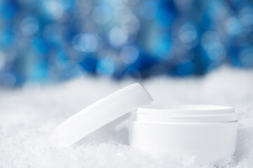 White jar of cream on the snow with a blurry shiny background. Blurred Christmas bokeh decor shines in blue. Dry skin care in winter. Cosmetic beauty product for face and hand skin care.