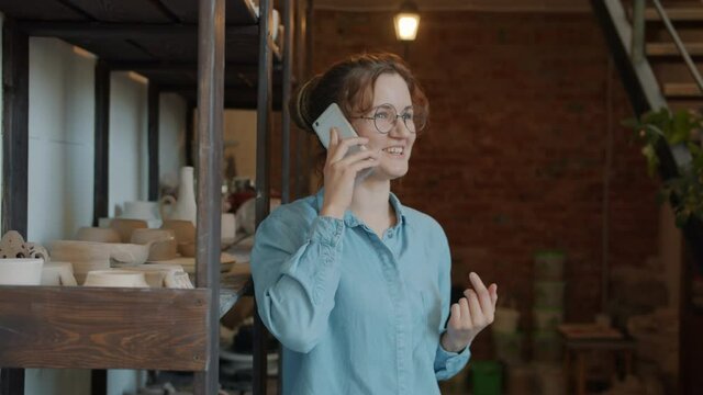 Happy young lady is speaking on mobile phone in pottery workshop smiling standing indoors alone. Youth, communication and modern technology concept.