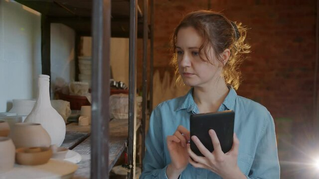 Slow motion of serious girl entrepreneur using tablet in pottery workshop looking at ceramics and touching device screen bucy with inventory.