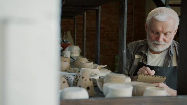 Portrait of old craftsman counting ceramics in workshop stock and writing in notebook busy with inventory. Family business and creative people concept.
