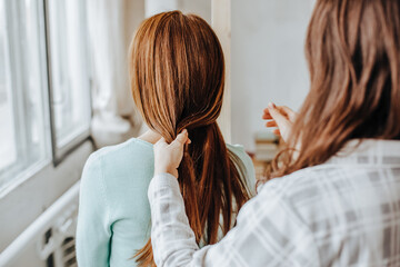 Two girls braid their hair at the window. Woman makes a braid to her friend. Hair weaving hairstyles. Girlfriend braids her hands with ringlets. Hair care
