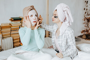 Two girls make masks and patches for the beauty of face and hair. Women take care of youthful skin. Girlfriends laugh, make faces and do beauty treatments at home with towels on their heads.