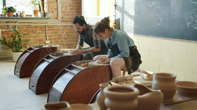 Slow motion of male and female ceramists working with pottery wheels in studio making earthenware and talking. Profession and hobby concept.