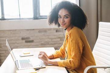 Attractive african american woman is using handfree headset for communication sitting at the desk with laptop at office modern space. Call center operator at work, sided view