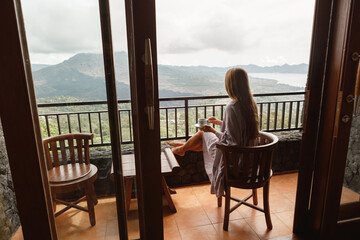 A young woman holding coffee cup while sittinhg on chair on balcony , looking at mountains and green nature