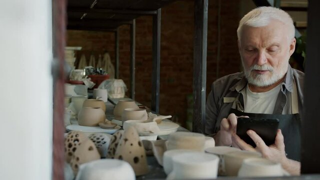 Portrait of senior craftsman counting hand-made pots in workshop and using tablet busy with inventory. Family business and technology concept.