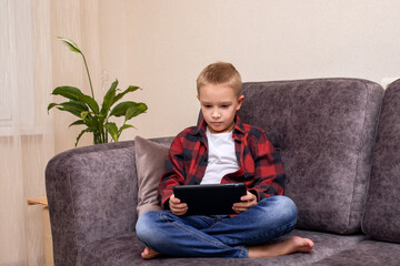 A charming 10-year-old boy in jeans and a checkered red shirt, wearing glasses and large headphones is listening to music or listening to a lesson on his smartphone