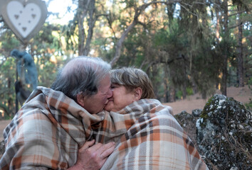 An affectionate kiss between two smiling elderly people in the woods. Around them many trees and a romantic wooden heart