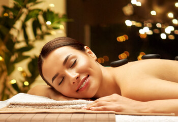 Obraz na płótnie Canvas wellness, beauty and relaxation concept - smiling beautiful young woman having hot stone massage at spa over festive lights on background