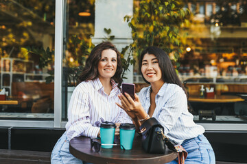 Smiling cheerful women friends drinking coffee using the phone looking at the camera sitting in a cafe on the street