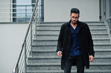 Middle eastern entrepreneur wear black coat and blue shirt, eyeglasses against office building smoking cigarette.