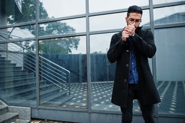 Middle eastern entrepreneur wear black coat and blue shirt, eyeglasses against office building smoking cigarette.