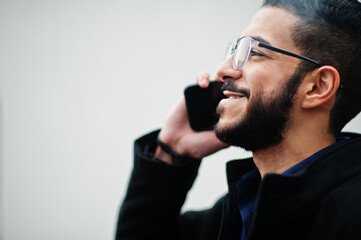 Middle eastern entrepreneur wear black coat and blue shirt, eyeglasses standing against white wall, smoking cigarette and speak by phone.