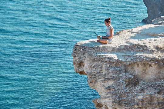 Girl using laptop for work. Sea and nature landscape from high cliff. Travelling and remote office concept, copy space
