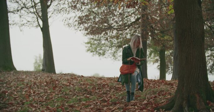 Young woman reading her diary to bring her memories back and to recover herself in a cold autumn journey on a swing under a big tree. slow motion