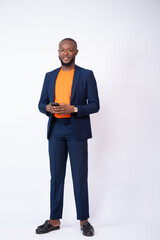 portrait of a young african man wearing a suit and holding his phone in texting and chatting manner smiling, standing against a white background