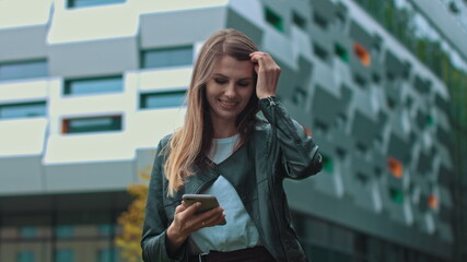 Good looking girl with Long Brown Hair Looking at Her phone Typing and Smiling. Enormous industrial Building at the Background. Green Bushes and Trees. Smart clothes. Natural makeup.