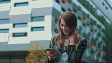 Good looking girl with Long Brown Hair Looking at Her phone Typing and Smiling. Enormous industrial Building at the Background. Green Bushes and Trees. Smart clothes. Natural makeup.