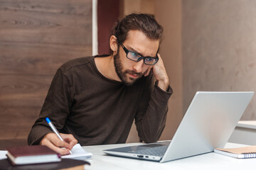 Serious young man with beard looks into laptop and writes in notepad