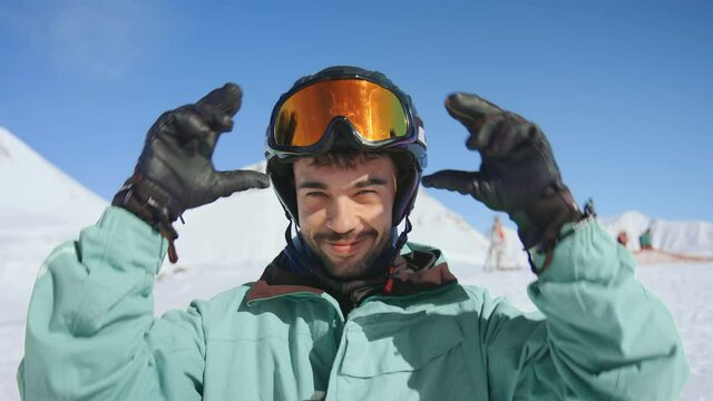 Young happy man looks into camera, smiles and squints in sun, excited to start extreme fast downhill on ski resort slope. Portrait of skier or snowboarder in winter outdoor outfit. Authentic hapiness