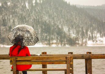 A view of alone and sad girl in red coat and black hair with umbrella sitting on a wooden bench  against the forest by the lake.