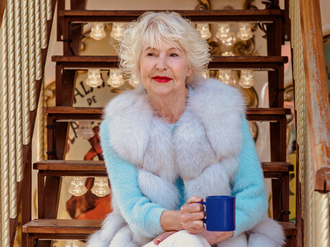 senior stylish woman in fur coat and with grey hair sitting on carousel drinking tea (coffee) and enjoying life. Travel, fun, happiness, seasonal concept