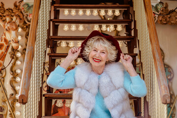 senior stylish woman in fur coat and with grey hair sitting on carousel holding purple hat. Travel, fun, fashion, happiness, seasonal concept © k8most