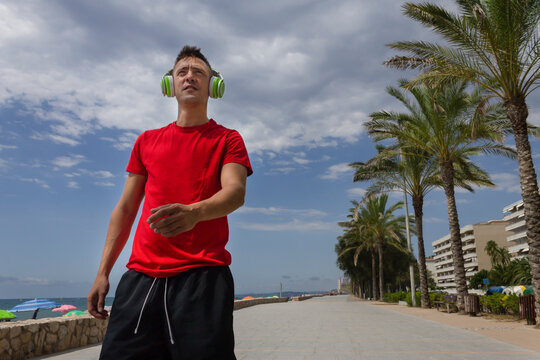 Handsome man wearing headphones listening music, chilling and walking on seafront promenade among palm trees.