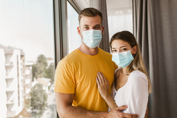 Young couple in isolation wearing mask standing by the window