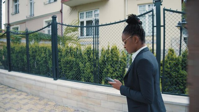 Black african woman entrepreneur walking in neighbourhood using smartphone ordering taxi to the airport. Businesswoman in smart-casual style. Mobile phone. Outdoors.