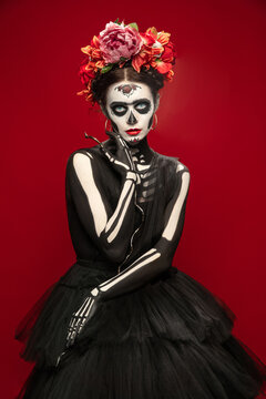 Scary. Young girl like Santa Muerte Saint death or Sugar skull with bright make-up. Portrait isolated on red studio background with copyspace. Celebrating Halloween or Day of the dead.