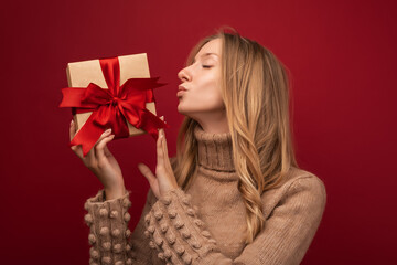 Charming blonde woman holding gift with red ribbon. Studio shot red background. New Year Birthday Holiday concept