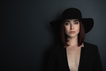 Fototapeta na wymiar Beautiful fashion woman with perfect body, female face. Portrait of charming girl wearing black blazer and hat posing in studio over deep gray background and looking at camera