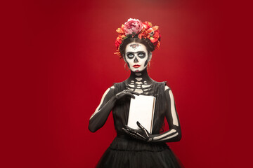 Book. Young girl like Santa Muerte Saint death or Sugar skull with bright make-up. Portrait isolated on red studio background. Celebrating Halloween or Day of the dead. Copyspace on cover.