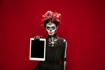 Holding tablet. Young girl like Santa Muerte Saint death or Sugar skull with bright make-up. Portrait isolated on red studio background. Celebrating Halloween or Day of the dead. Copyspace on screen.